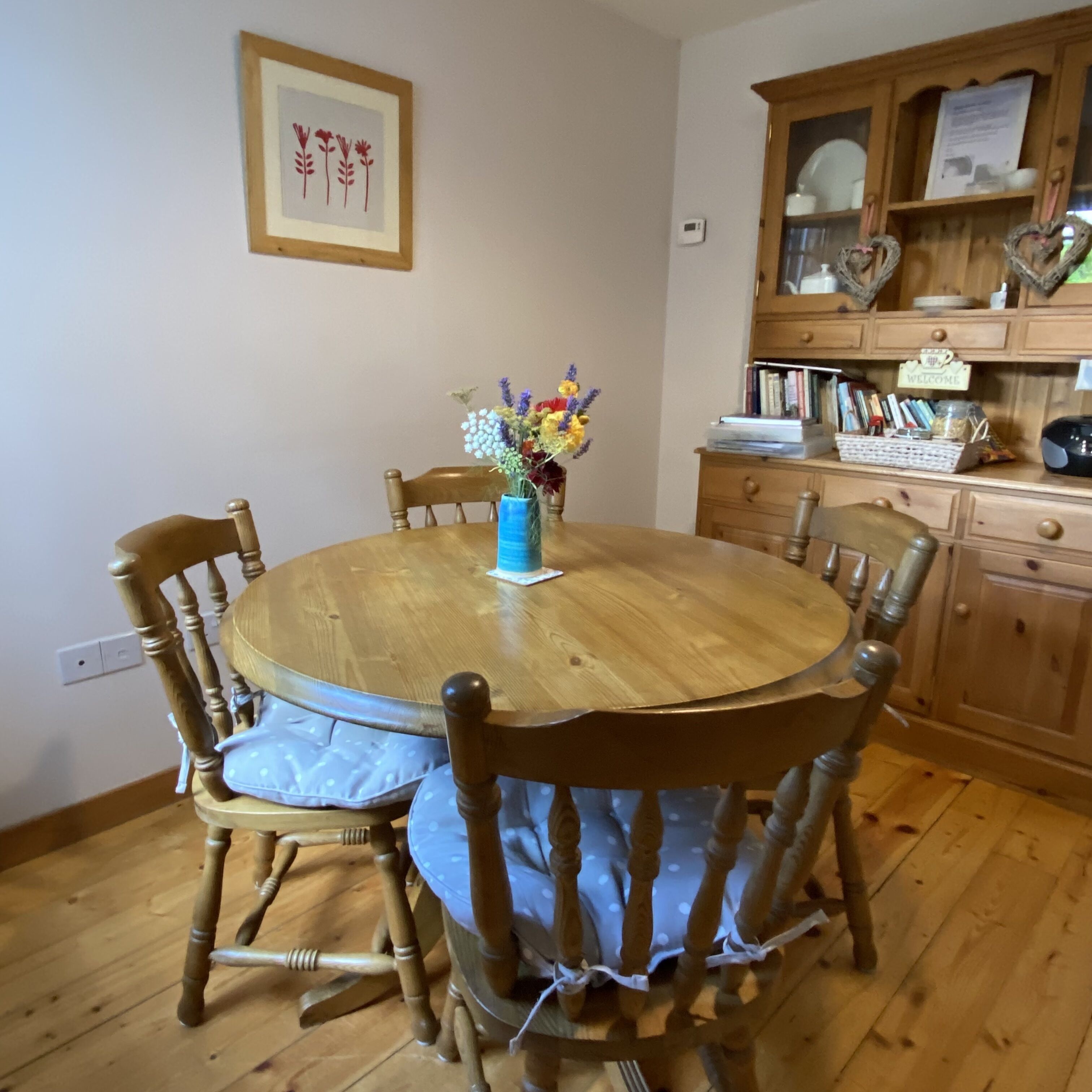 A round wooden dining table with six chairs, a vase with flowers, and a wooden cabinet with books and a welcome sign.