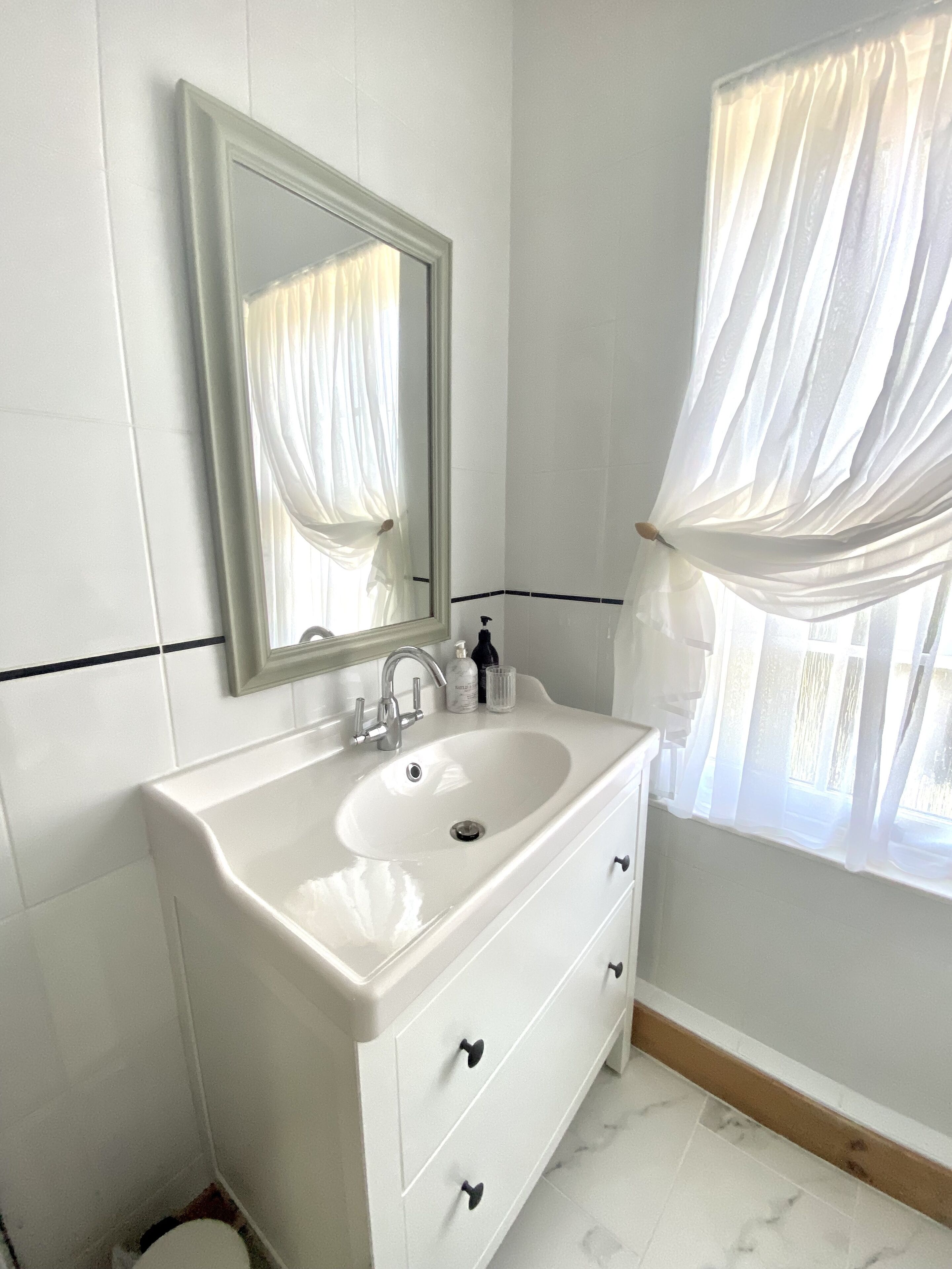 A bathroom with a white sink, a mirror, and a window with sheer curtains.