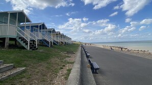 A row of beach huts with steps leading up to them, a paved path beside the beach, and the ocean in the background. - Green Cottages (Sittingbourne)