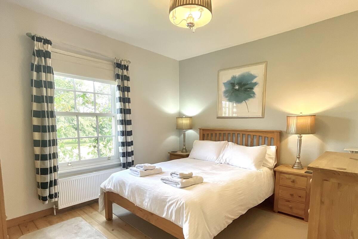 A bedroom with a wooden bed, a window with curtains, a bedside table with a lamp, and a radiator.