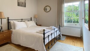 A bedroom with a bed, bedside lamps, a chandelier, a window with curtains, and a radiator. - Green Cottages (Sittingbourne)