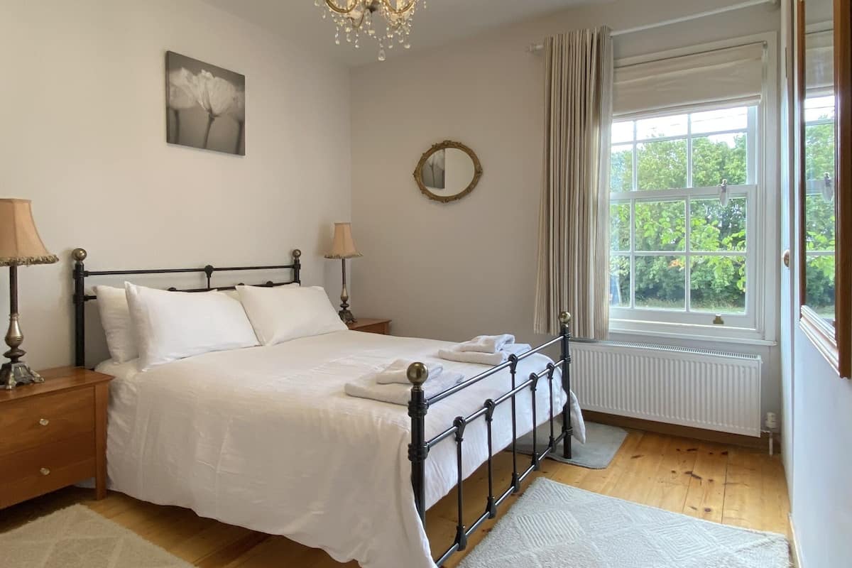 A bedroom with a bed, bedside lamps, a chandelier, a window with curtains, and a radiator.