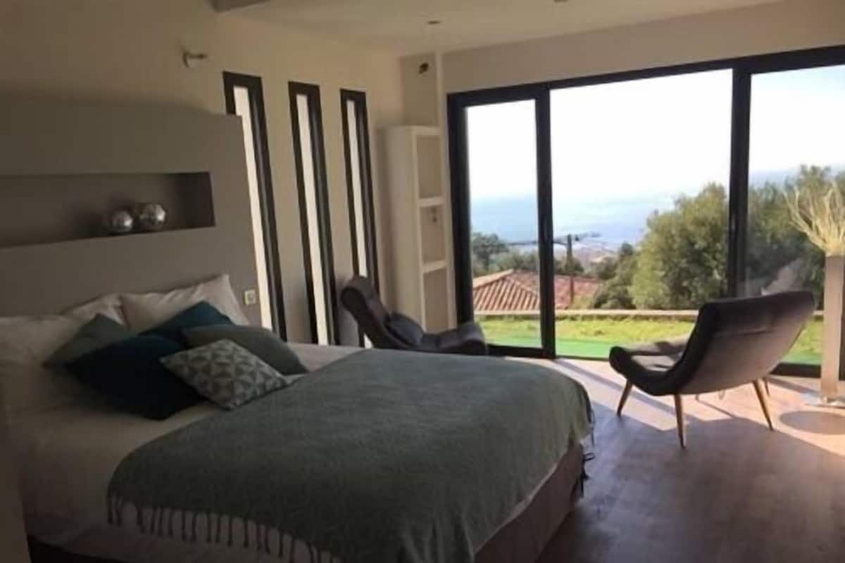 A modern bedroom with a large bed, a chair, and a view of the outdoors.