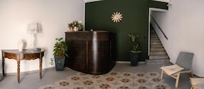 Un área de recepción con un mostrador de madera oscura, una mesita con una lámpara y una silla. Hay plantas en macetas y un adorno en la pared.