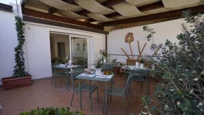 Un patio exterior con mesas y sillas, plantas en macetas y una puerta que da a un espacio interior.