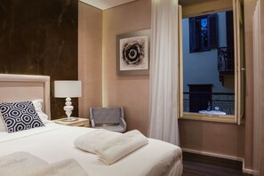 A hotel room with a bed, a nightstand, a lamp, a chair, a window with a view of the outside, and a framed artwork on the wall. - Consolato Boutique (Chania)