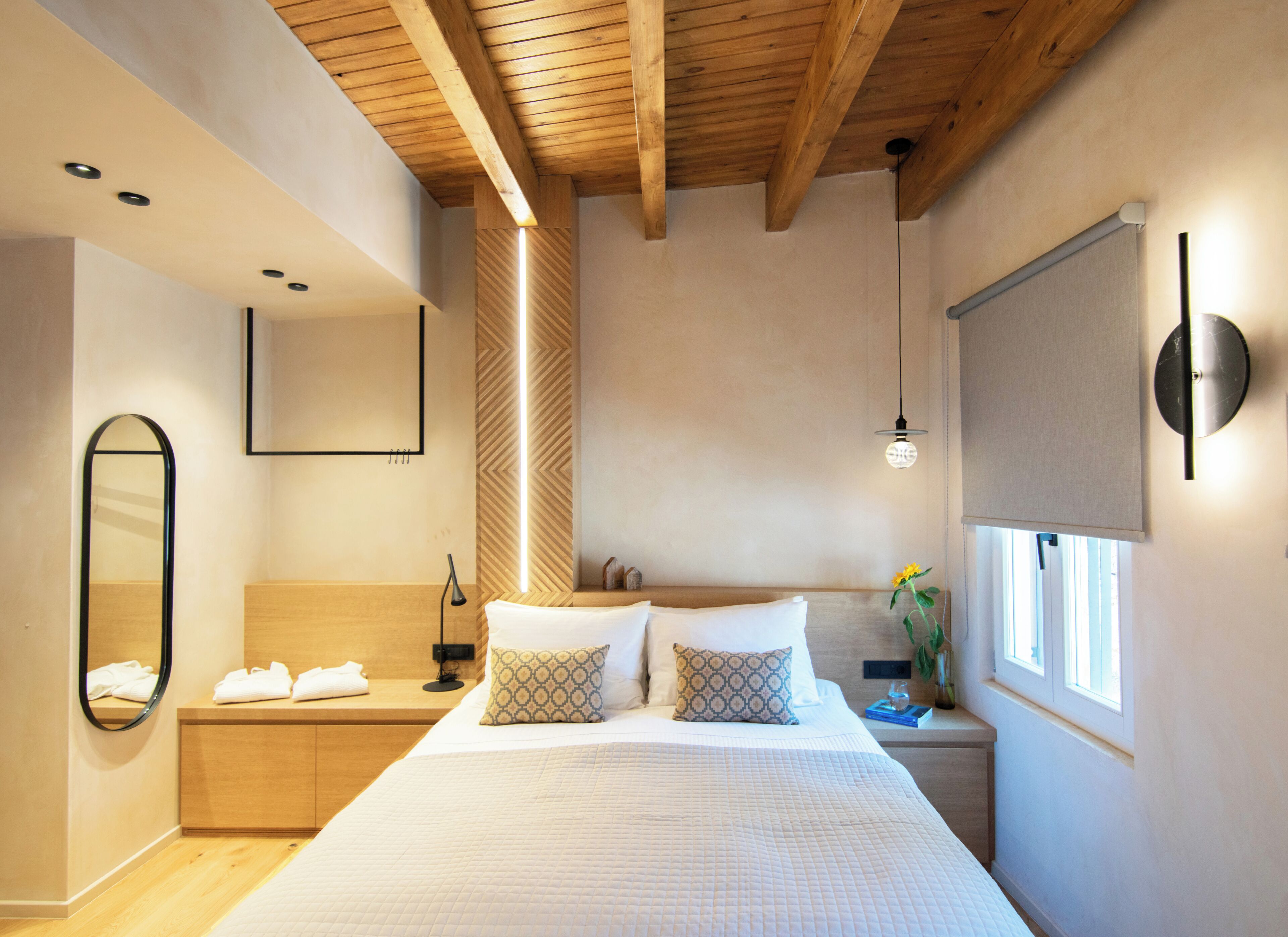 Een moderne slaapkamer met een groot bed, houten balken in het plafond, een nachtkastje met een lamp, een spiegel en een raam met zonwering.