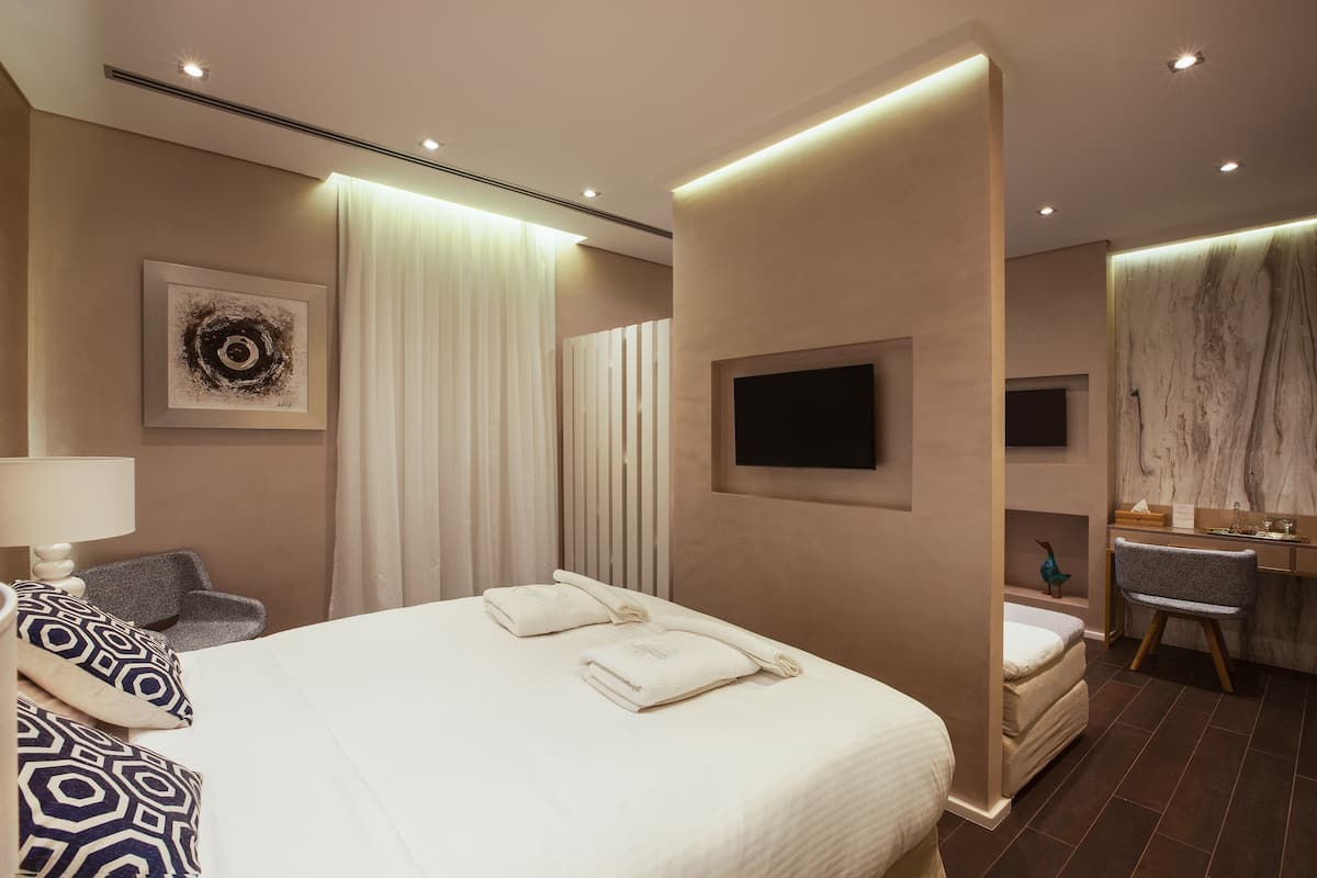 A modern hotel room with a large bed, a desk with a lamp, a chair, a TV mounted on the wall, and a bathroom with a mirror and sink.
