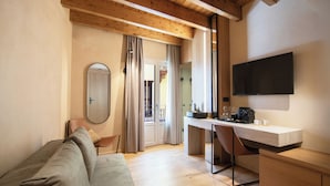 A modern room with a wooden ceiling, a sofa, a desk with a chair, a TV mounted on the wall, and a window with curtains. - Consolato Boutique (Chania)