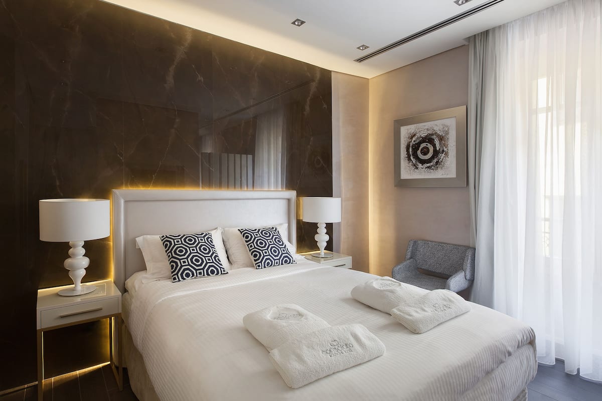 A modern hotel room with a large bed, two bedside tables with lamps, a gray armchair, and a painting on the wall.