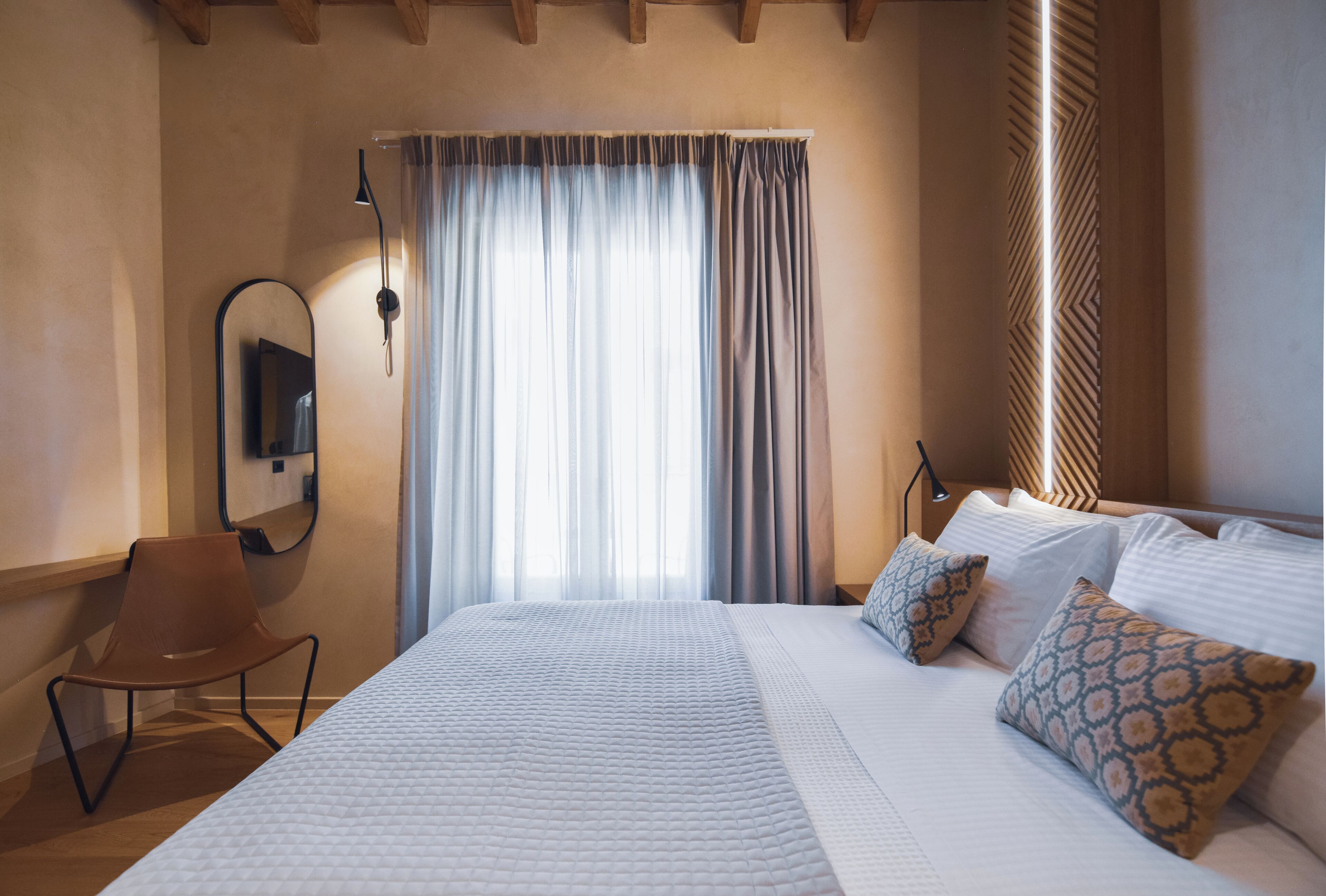 Une chambre d’hôtel avec un lit, une chaise, un miroir et une fenêtre avec des rideaux.