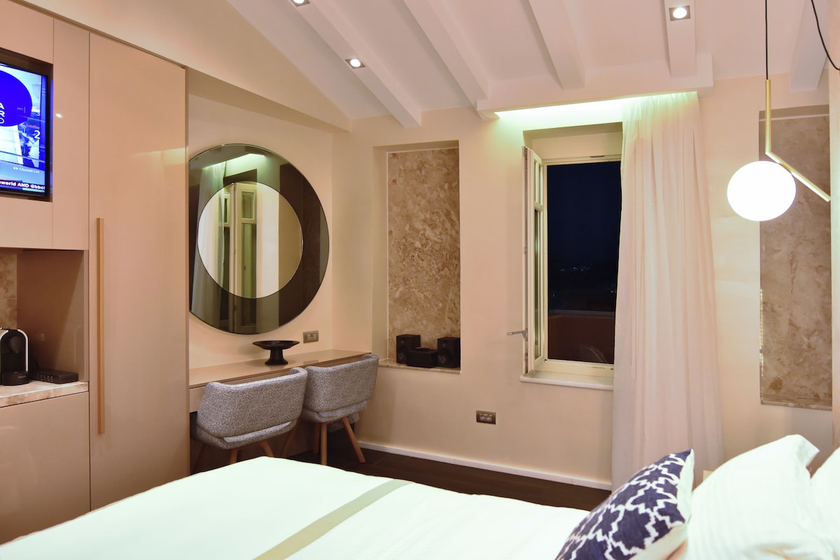A hotel room with a bed, a desk with two chairs, a round mirror, a television, and a window with curtains.
