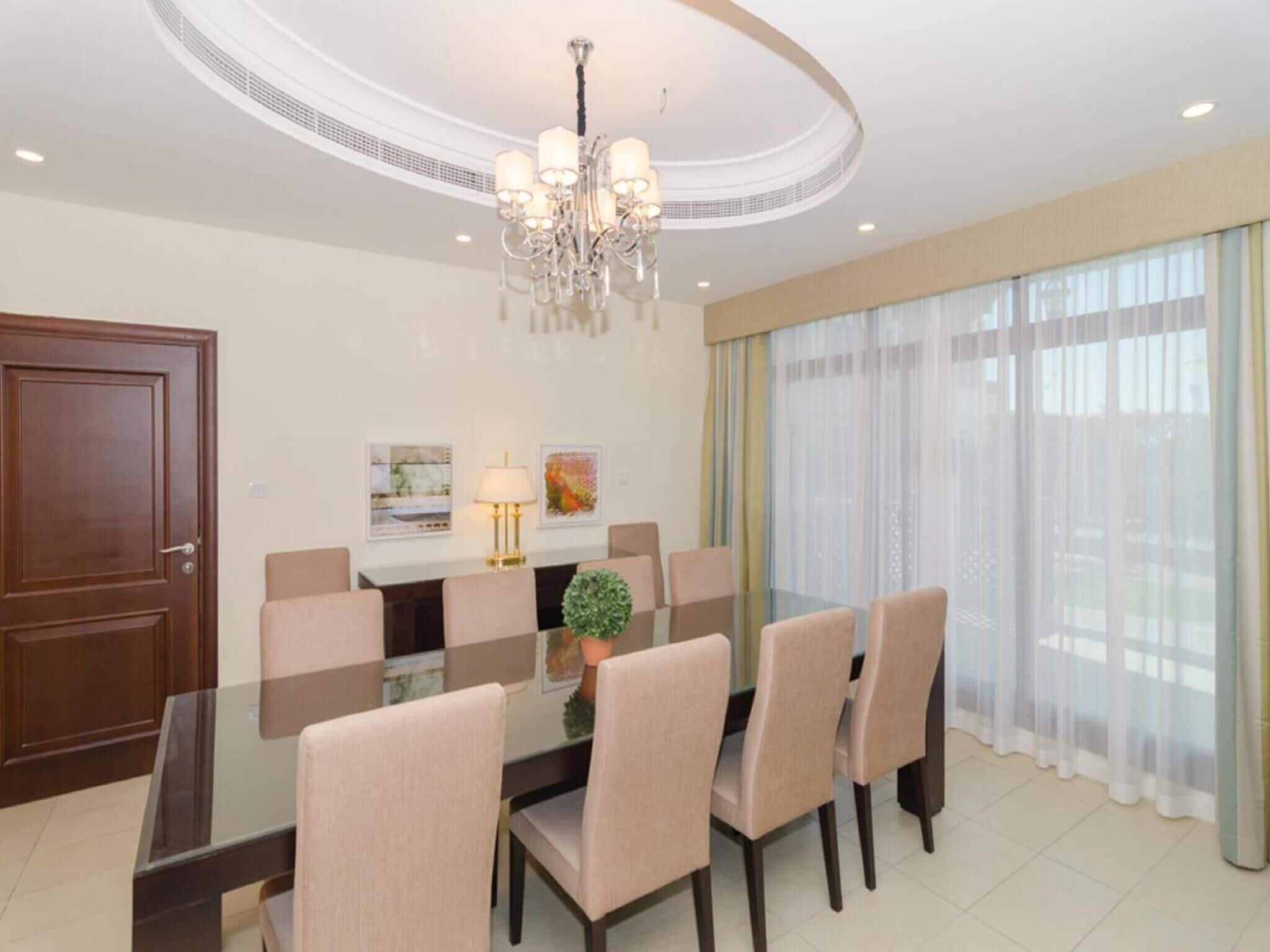 A modern dining room with a glass table, beige chairs, a chandelier, and a large window with sheer curtains.