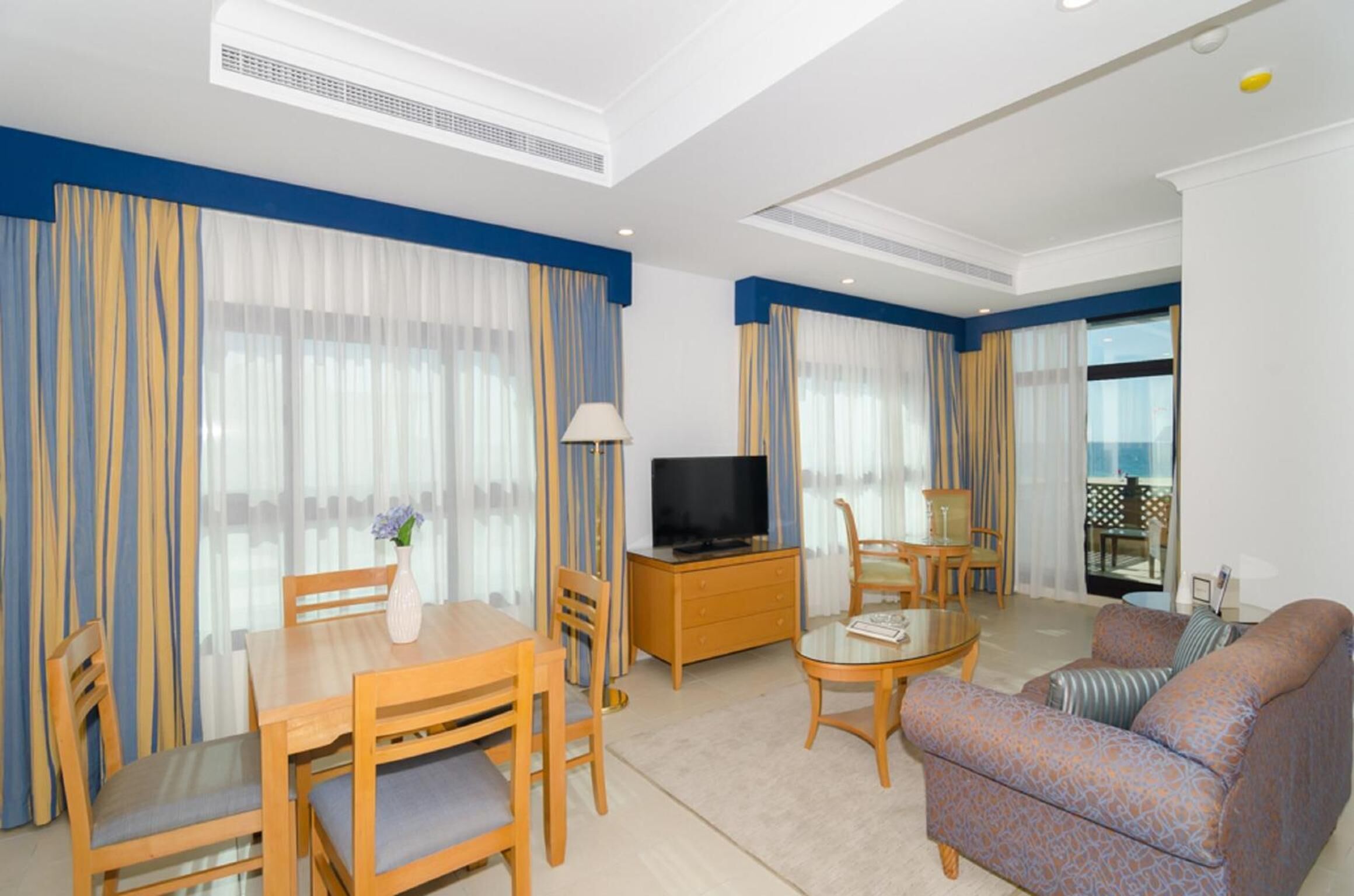 Habitación de hotel con zona de comedor, sofá, televisor y balcón con mesa y sillas.