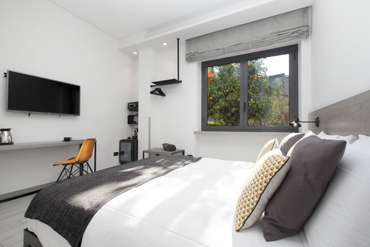 Une chambre moderne avec un grand lit, un téléviseur fixé au mur, un bureau avec une chaise et une fenêtre donnant sur des arbres.