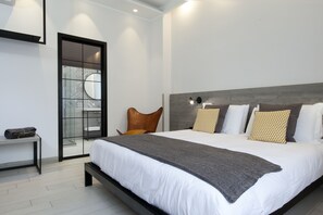 A modern bedroom with a large bed, a bedside table, a chair, and a glass door leading to a bathroom.