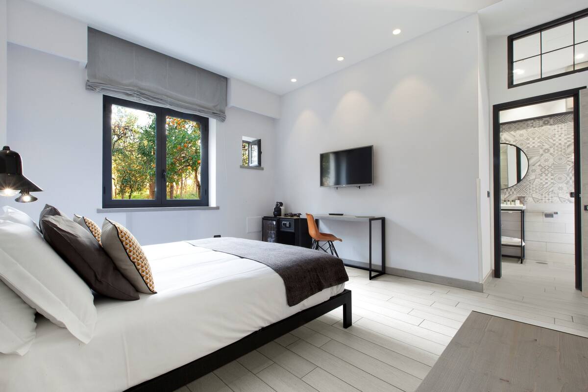Une chambre moderne avec un lit, un bureau, une télévision et une salle de bain visible à travers une porte ouverte.