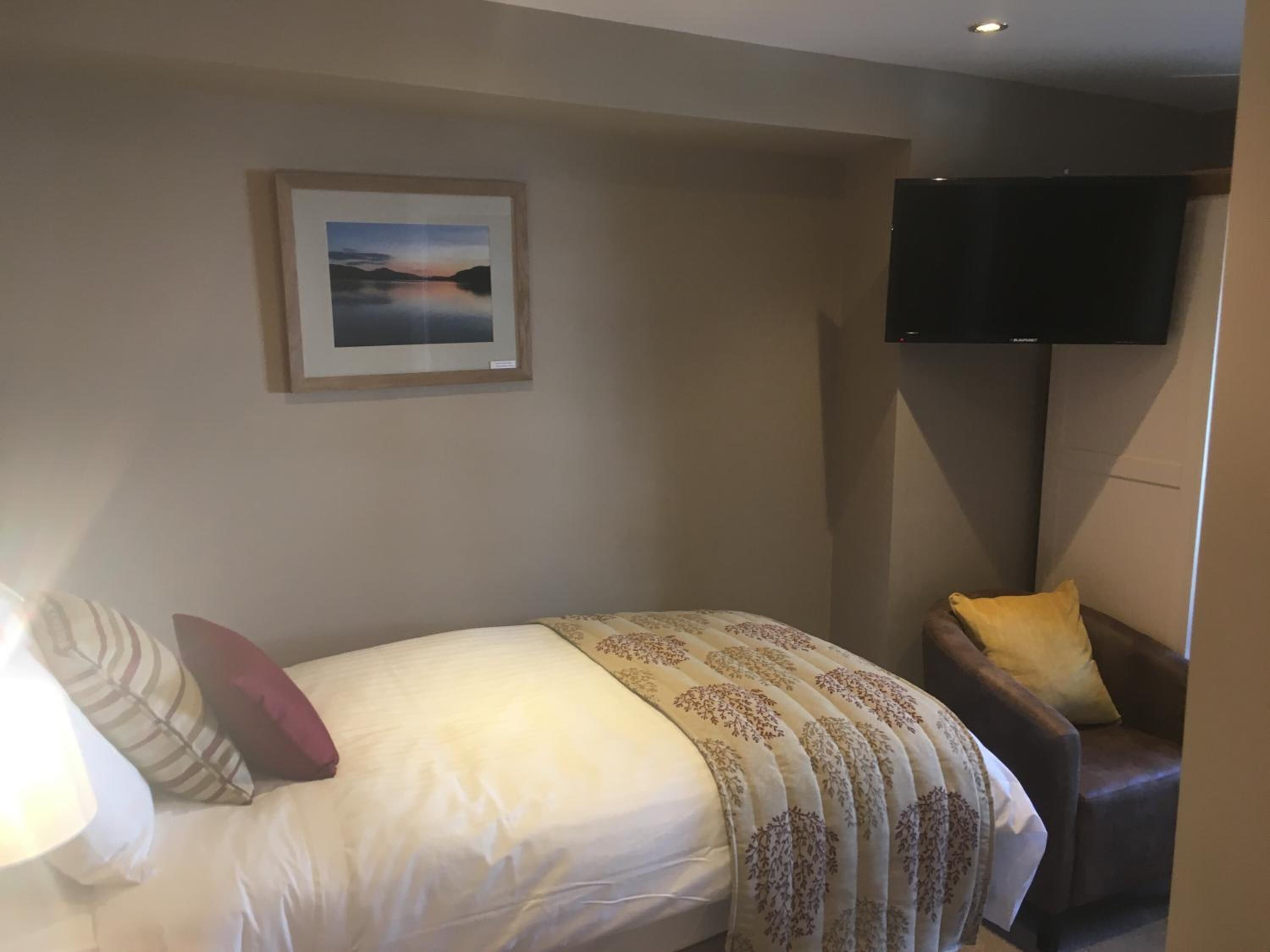 Une chambre d’hôtel avec un lit, une chaise, un téléviseur à écran plat fixé au mur et un tableau encadré au-dessus du lit.