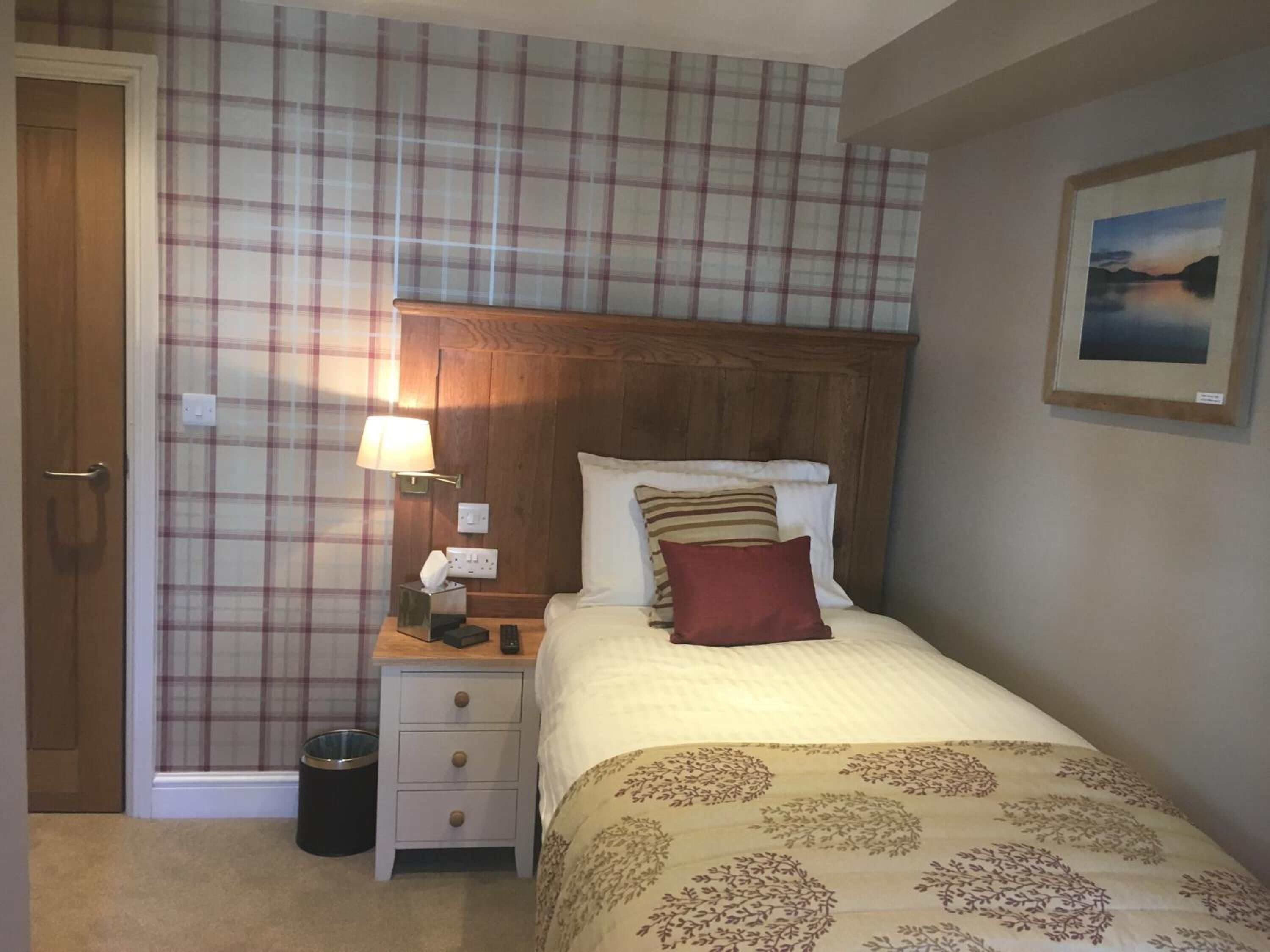 A neatly arranged bedroom with a plaid-patterned wall, a wooden headboard, a bedside table with a lamp, a trash bin, and a framed picture on the wall.