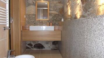 A bathroom with a stone wall, a wooden vanity, a white sink, a mirror, and a bidet.