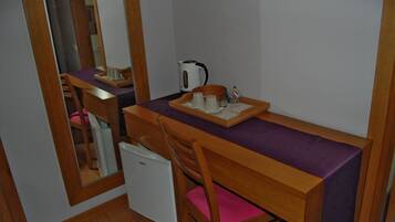 A compact room with a wooden desk, a chair, a small refrigerator, a kettle, and a mirror.