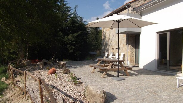 A patio area with a wooden table and chairs, a white umbrella, and a stone wall. - Quinta dos Caibrais (Oliveira de Frades)