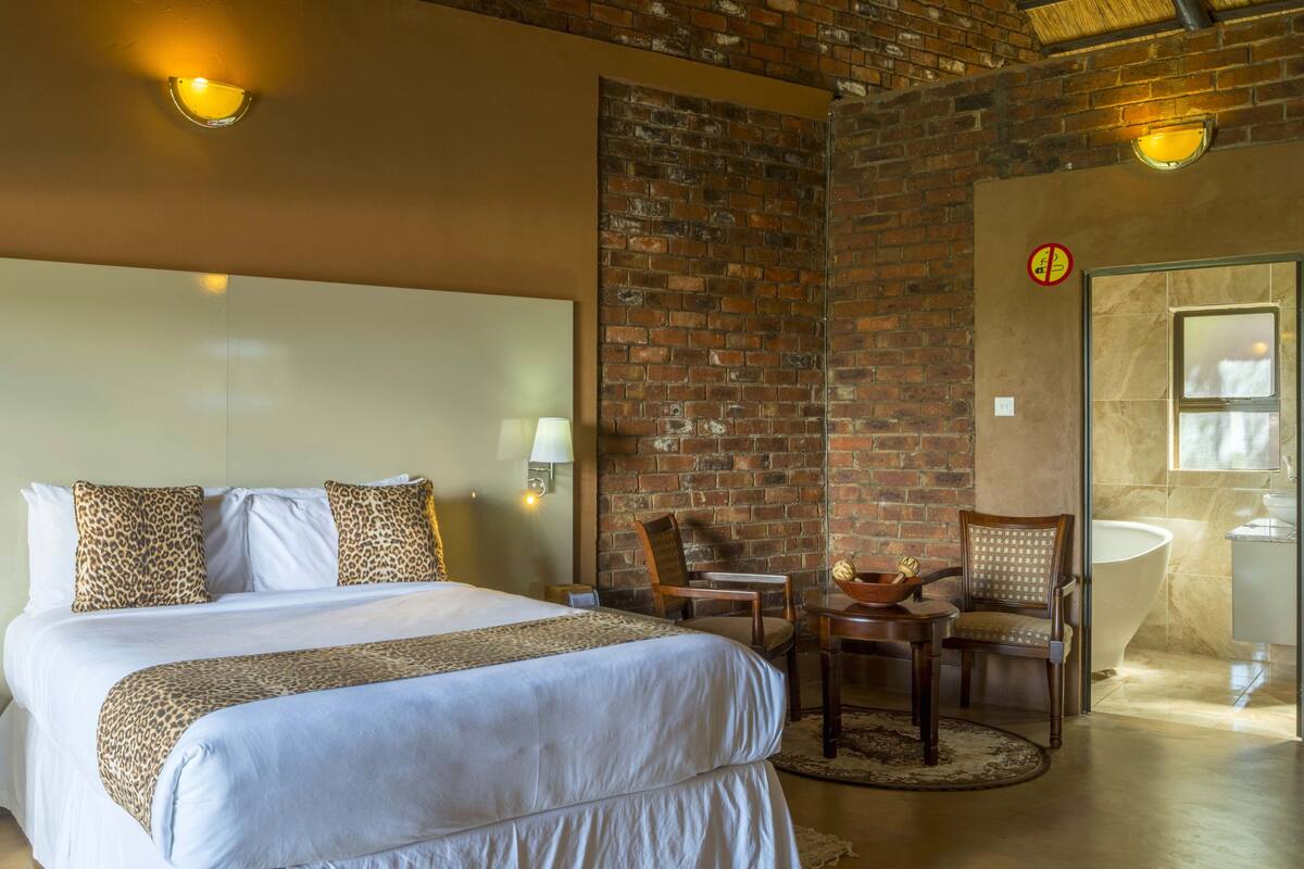 A hotel room with a large bed, two bedside lamps, a brick wall, a wooden chair, a small table with a bowl, and a bathroom with a bathtub and sink.