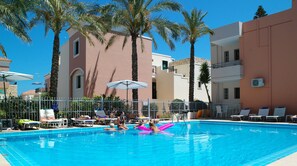 A swimming pool with lounge chairs and umbrellas, surrounded by palm trees and buildings. - Kallitsakis Beach (Platanias)