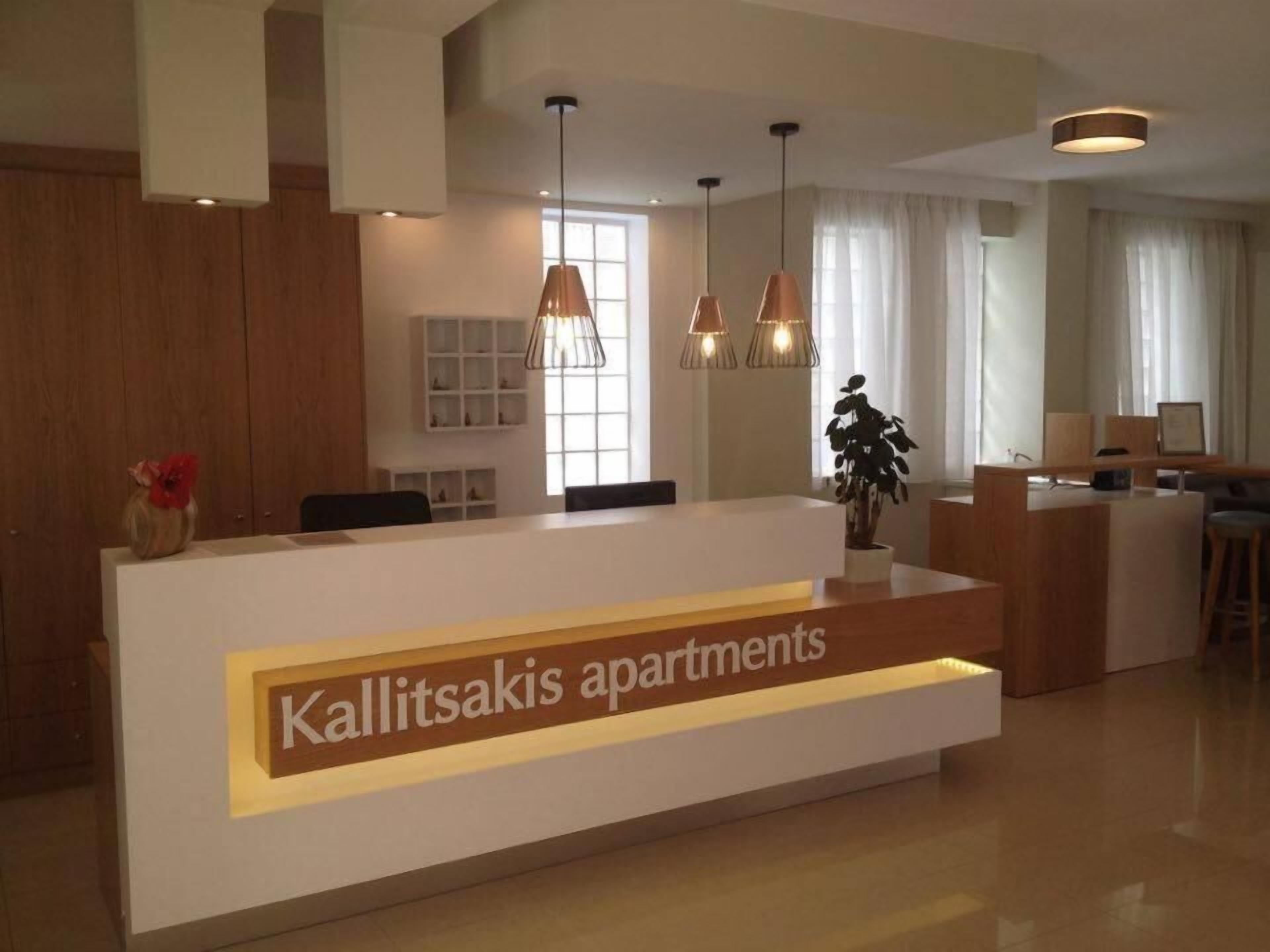 A reception area with a modern design, featuring a reception desk labeled "Kallitsakis apartments" and a large window providing natural light.