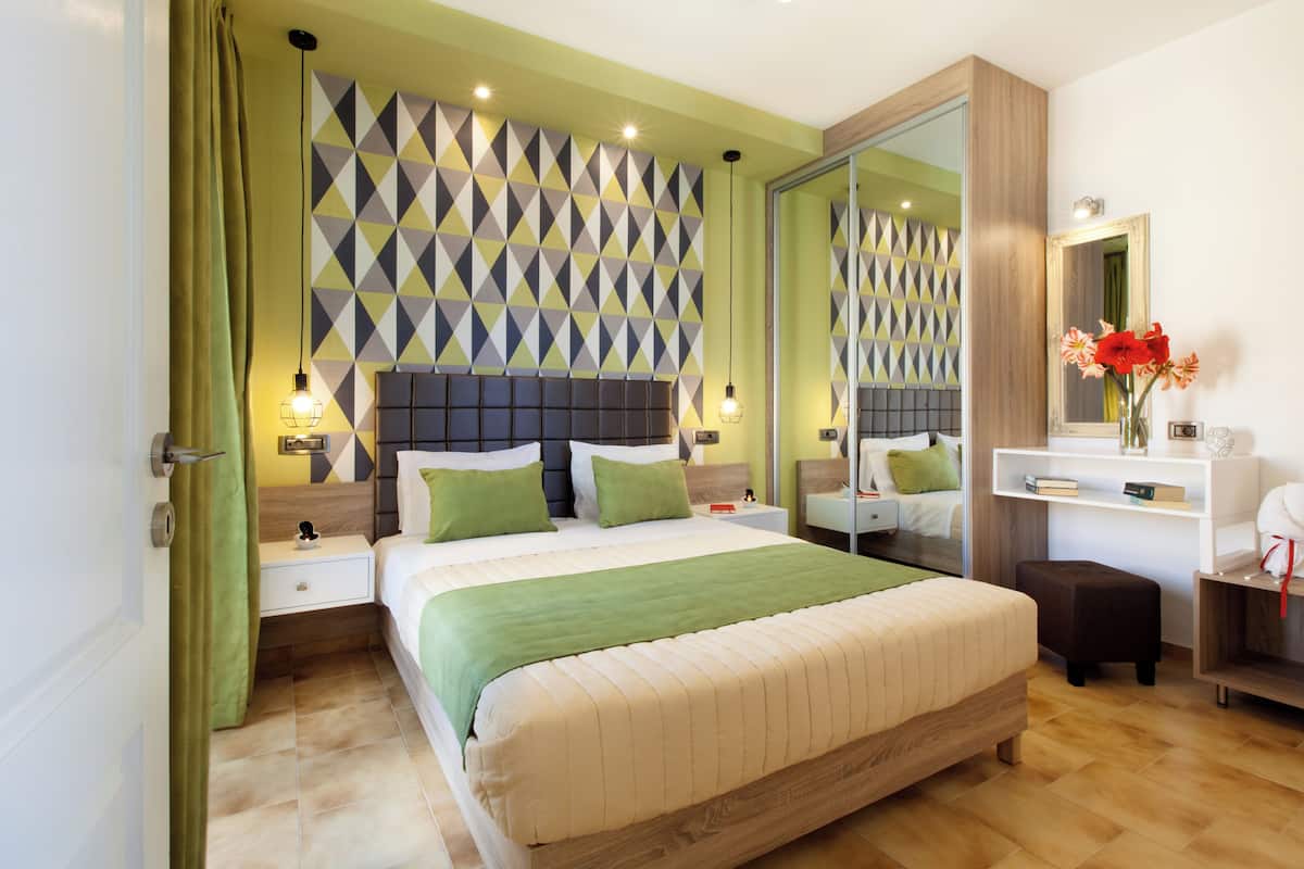 A modern bedroom with a large bed, green bedding, a geometric-patterned headboard, bedside tables with lamps, a mirror, and a small desk with a vase of flowers.