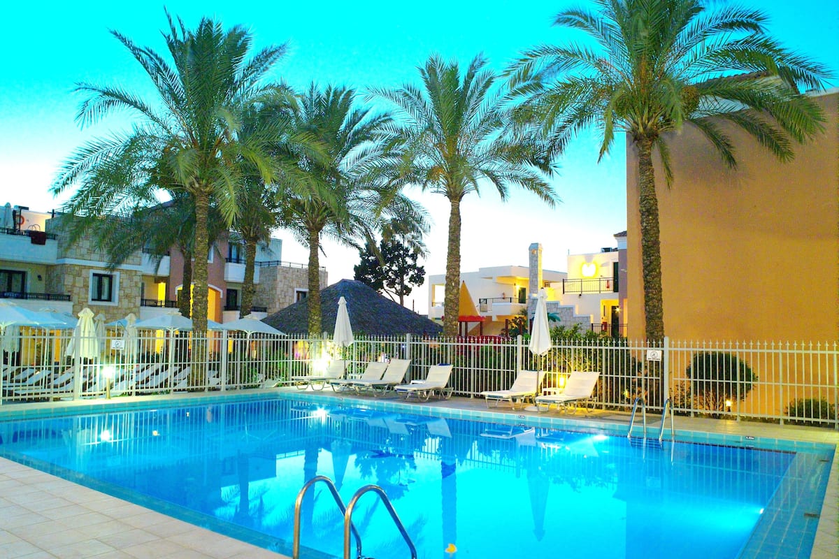 A swimming pool with lounge chairs and umbrellas, surrounded by palm trees and a building with a mountain-like structure in the background.
