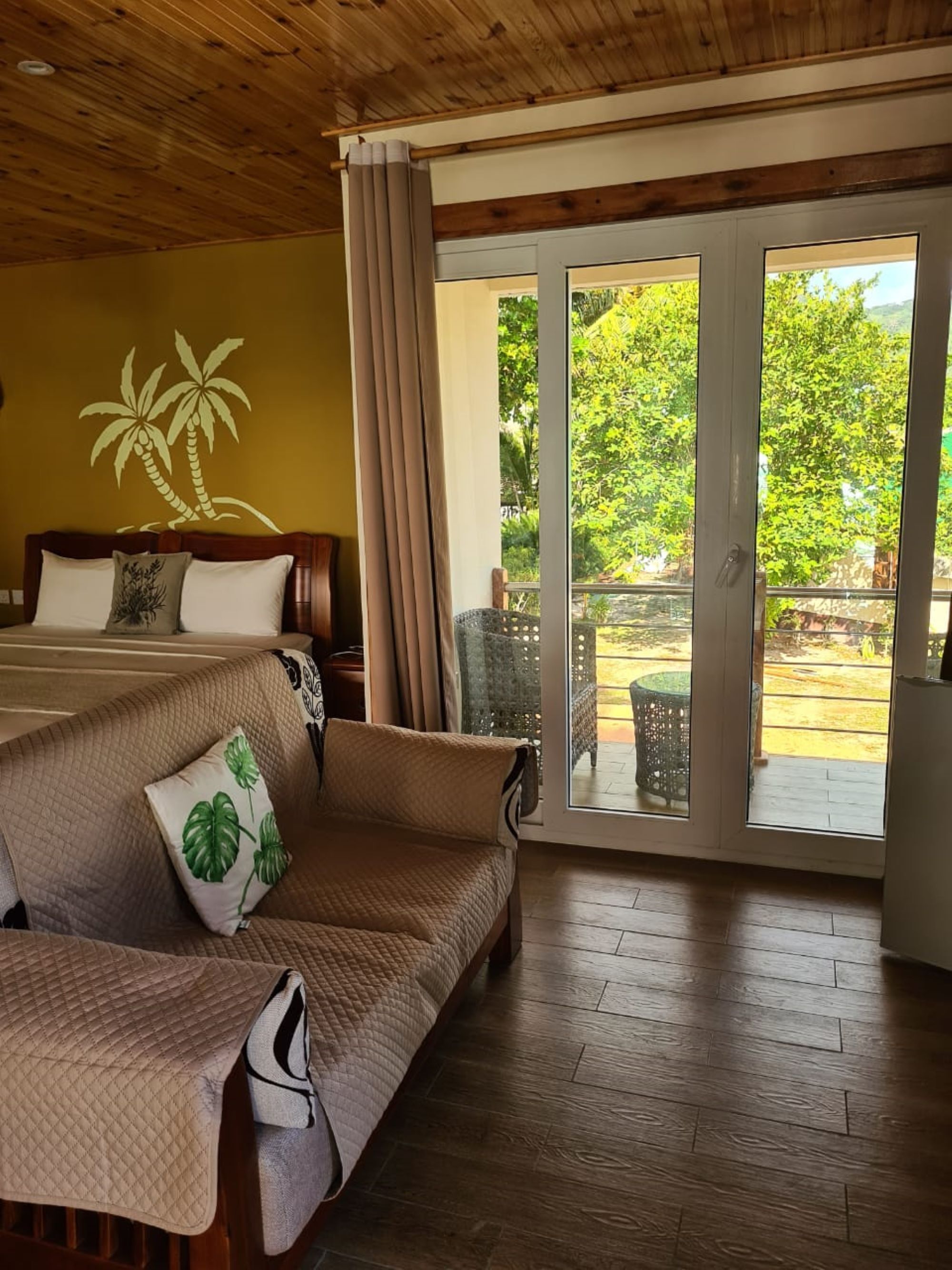 A room with a bed, a sofa, and a sliding glass door leading to an outdoor area with greenery.