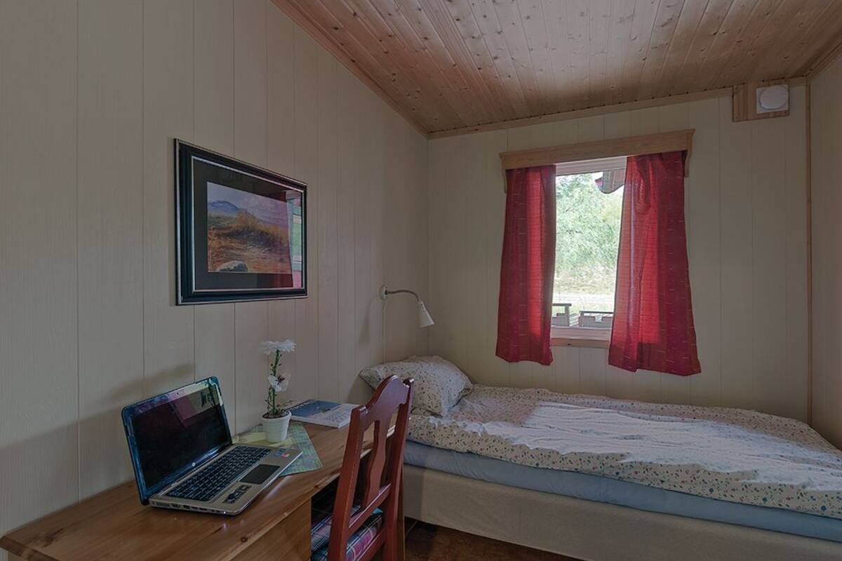 A small room with a wooden desk, a laptop, a bed, a window with red curtains, and a framed picture on the wall.