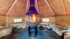 A wooden cabin interior with a central wood-burning stove, fur-covered benches, and a high wooden ceiling.