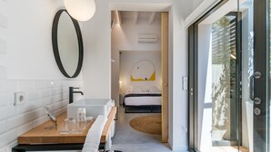 Een moderne badkamer met een spiegel, wastafel en handdoekhouder. Achter een open deur is een slaapkamer zichtbaar, met een bed en een rond vloerkleed.