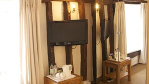 A room with exposed wooden beams, a television mounted on the wall, a small wooden table with a cup and bottles, and a mirror reflecting the room. - Chantry Hotel (Bury St Edmunds)