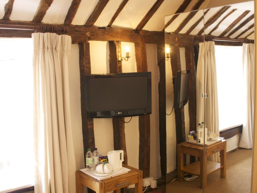 A room with exposed wooden beams, a television mounted on the wall, a small wooden table with a cup and bottles, and a mirror reflecting the room.