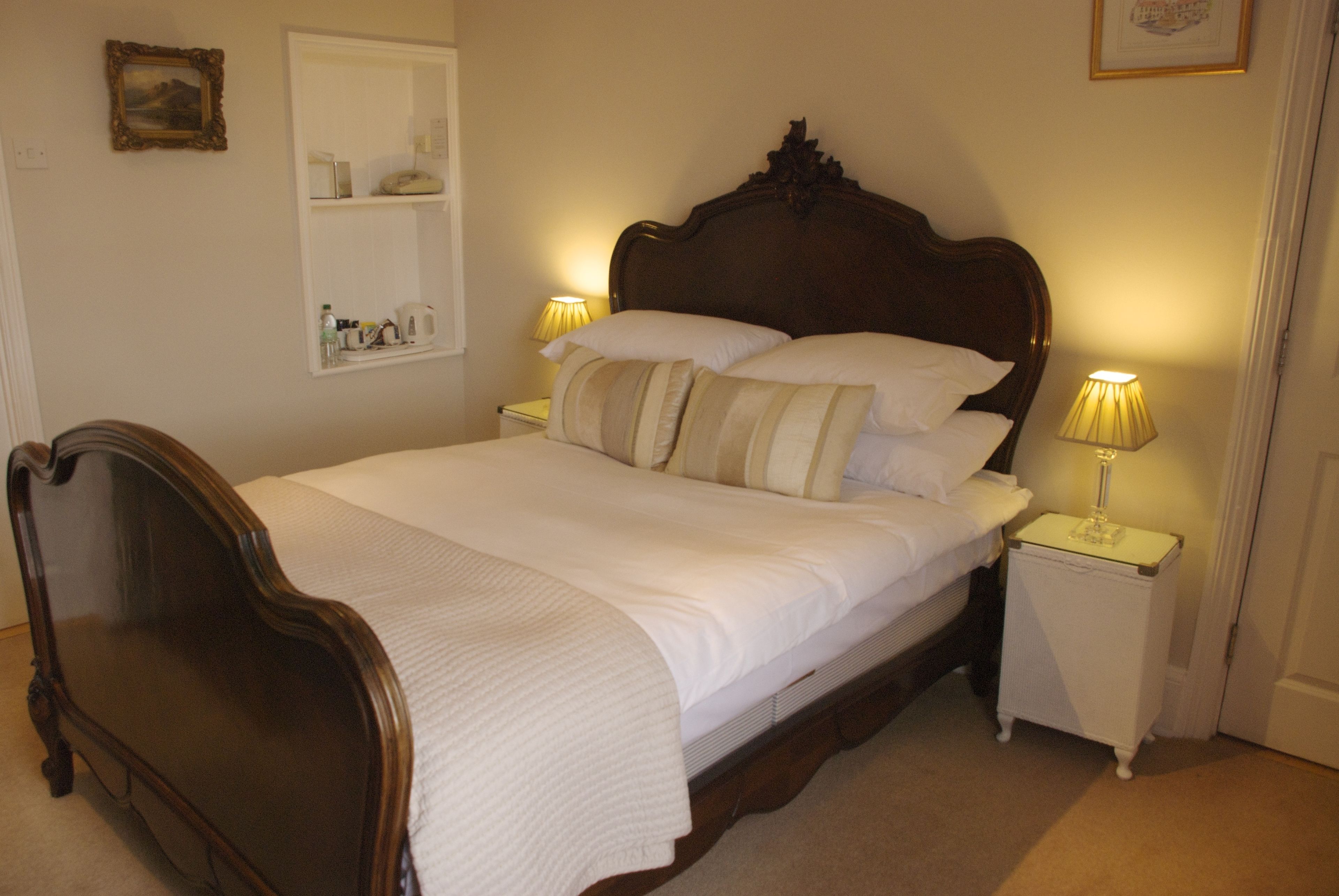 A neatly made bed with a wooden headboard, two bedside tables with lamps, a shelf with toiletries, and a framed picture on the wall.
