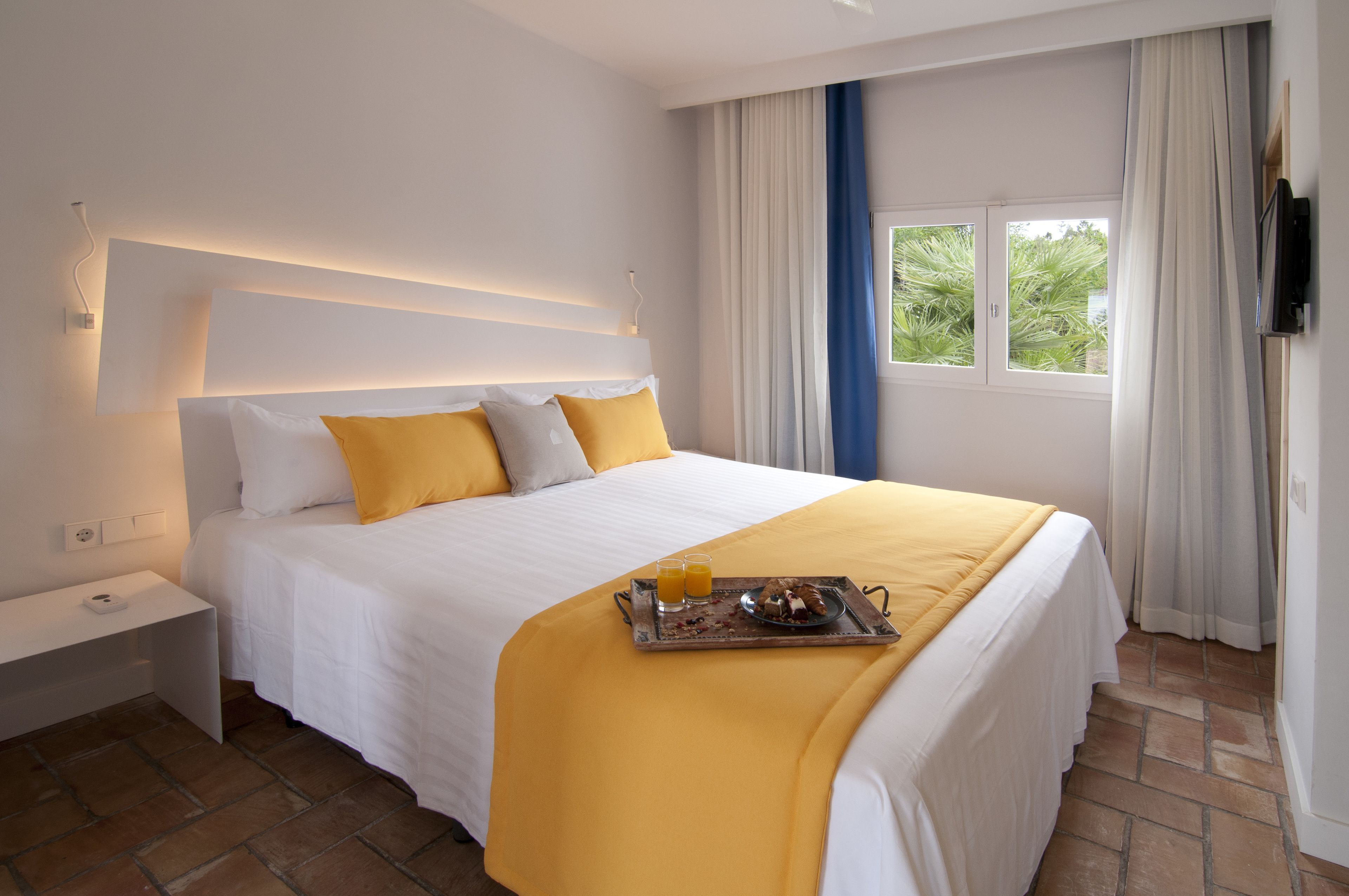 A neatly made bed with a tray of breakfast, a bedside table, and a window with curtains.