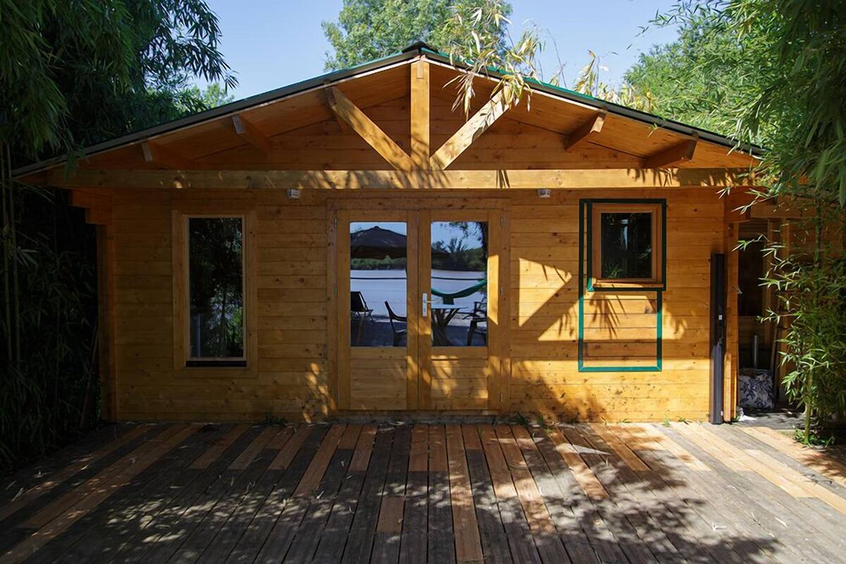 A wooden cabin with a deck, large windows, and a view of the water.