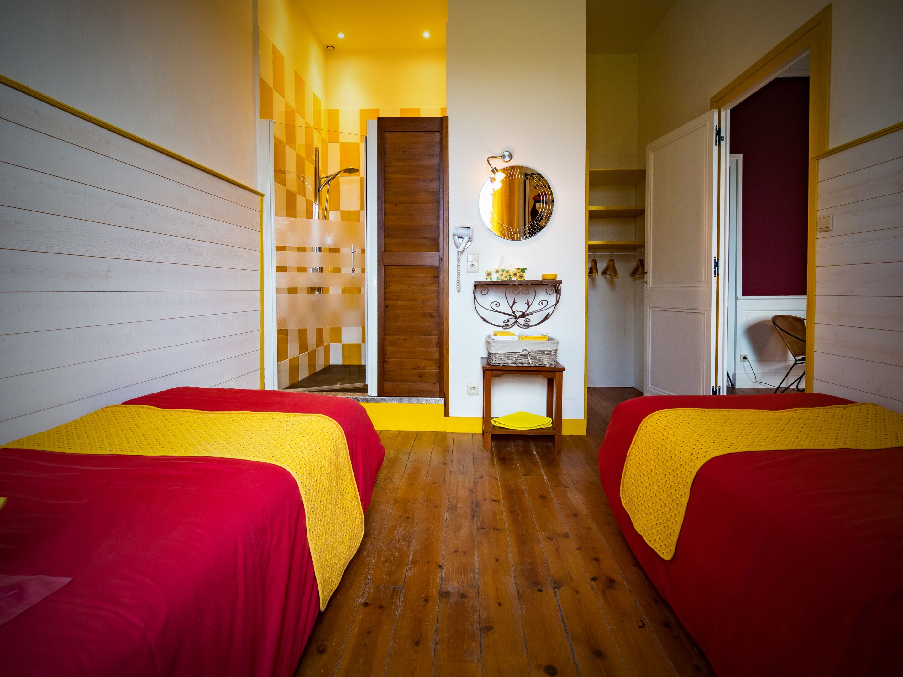 Une chambre d’hôtel avec deux lits simples, un plancher en bois, une salle de bain avec douche, un miroir et une petite table avec un panier.