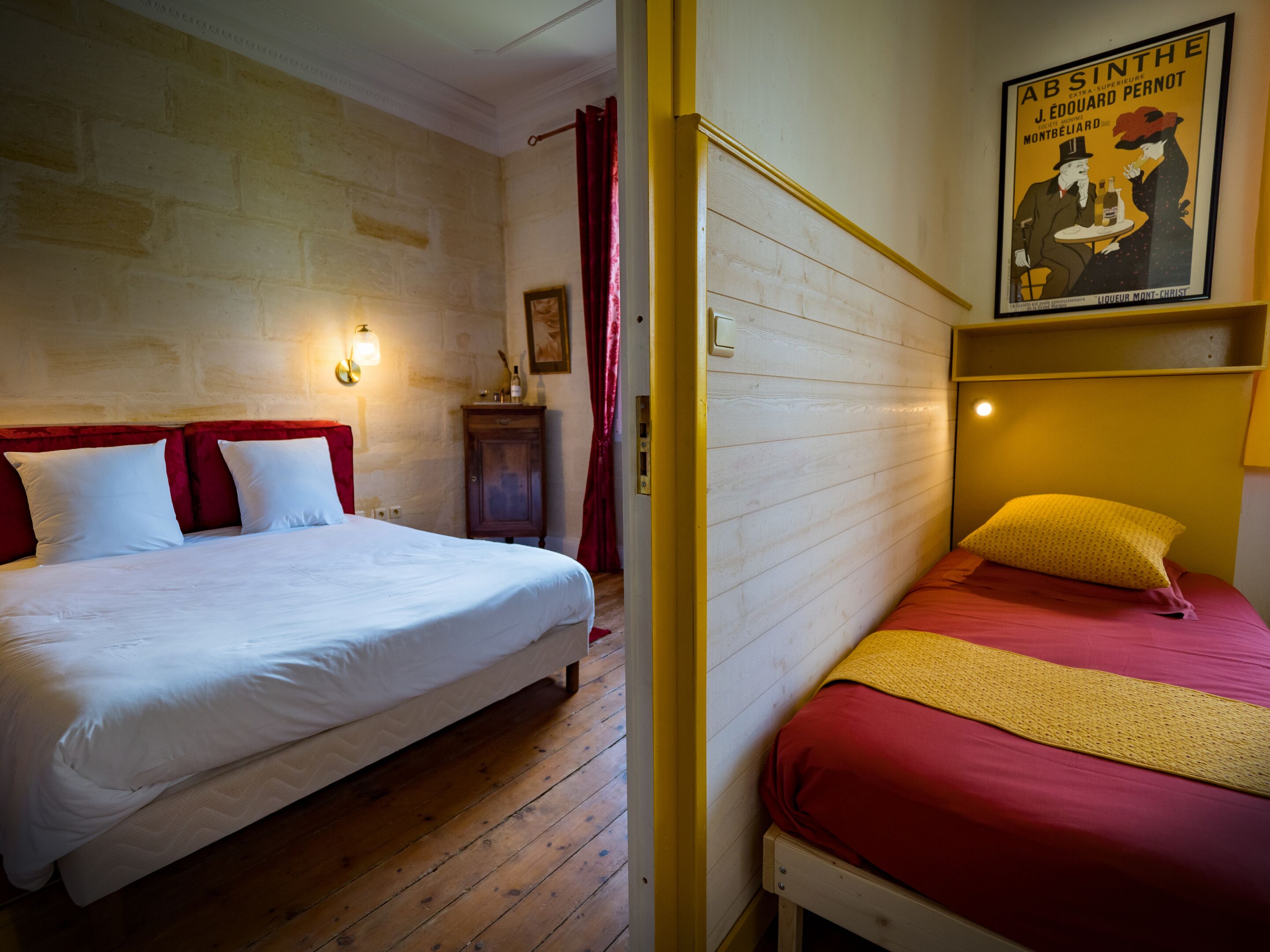 Una habitación con dos camas, una con ropa de cama blanca y la otra con ropa de cama roja. El suelo es de madera, hay un interruptor de luz en la pared y un cartel en la pared.