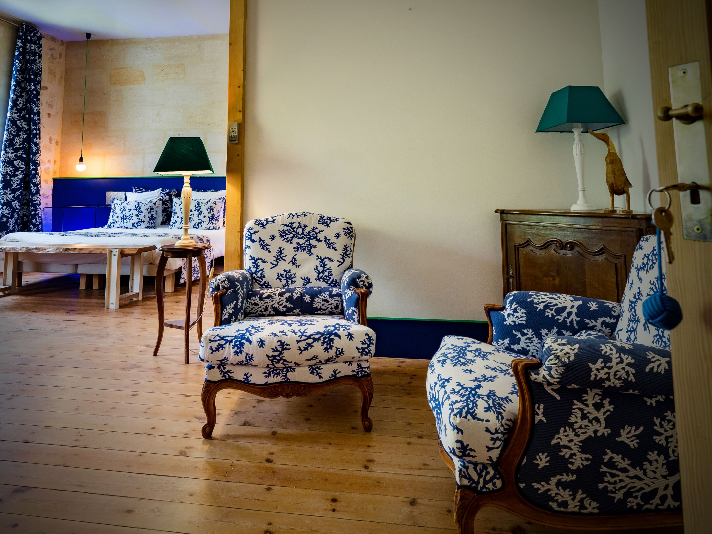 A room with a wooden floor, two blue and white patterned armchairs, a wooden cabinet, a green lamp, and a bed with matching bedding.