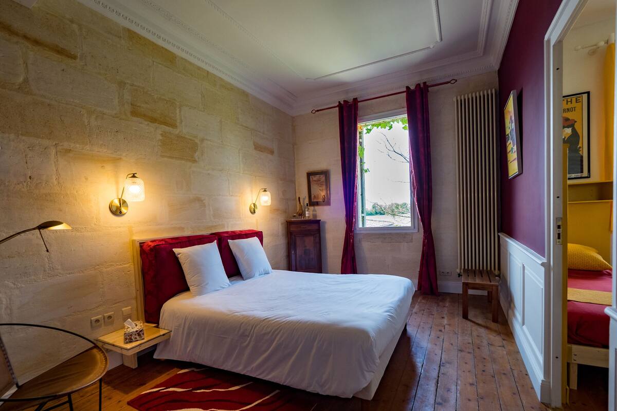 A bedroom with a large bed, a nightstand, a chair, a window with curtains, and a radiator.
