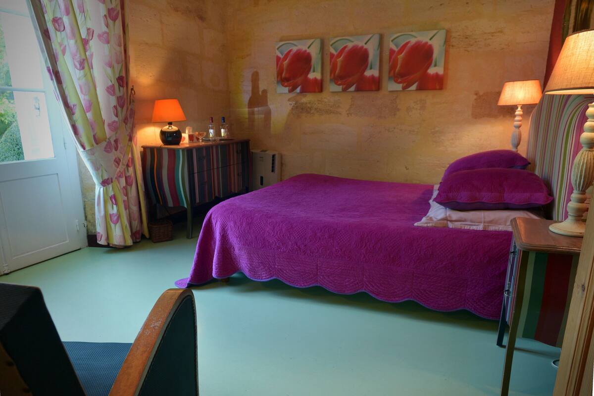 A bedroom with a purple bedspread, a striped side table, and a lamp. There are three paintings on the wall and a window with curtains.