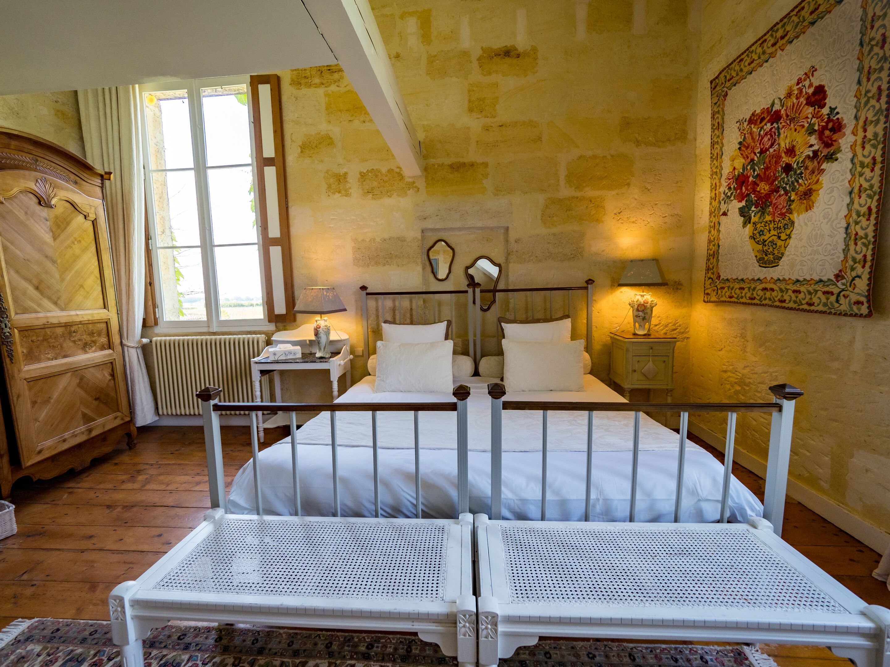 A room with two single beds, a wooden wardrobe, a radiator, a window with curtains, and a floral tapestry on the wall.