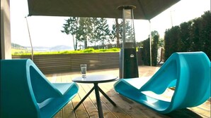 A covered outdoor seating area with two blue chairs and a small table, overlooking a landscaped garden and distant hills.