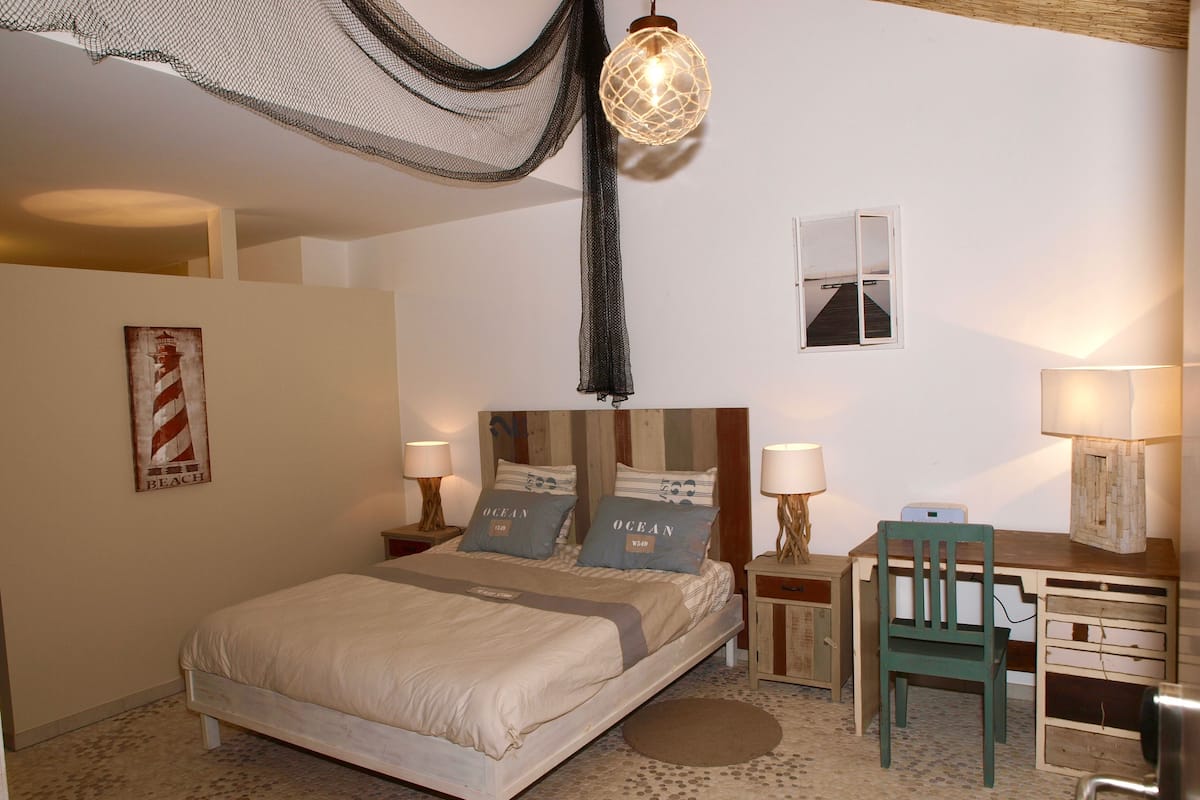 Une chambre à coucher avec un lit, deux tables de chevet avec des lampes, un bureau avec une chaise, un tableau accroché au mur et un luminaire suspendu.