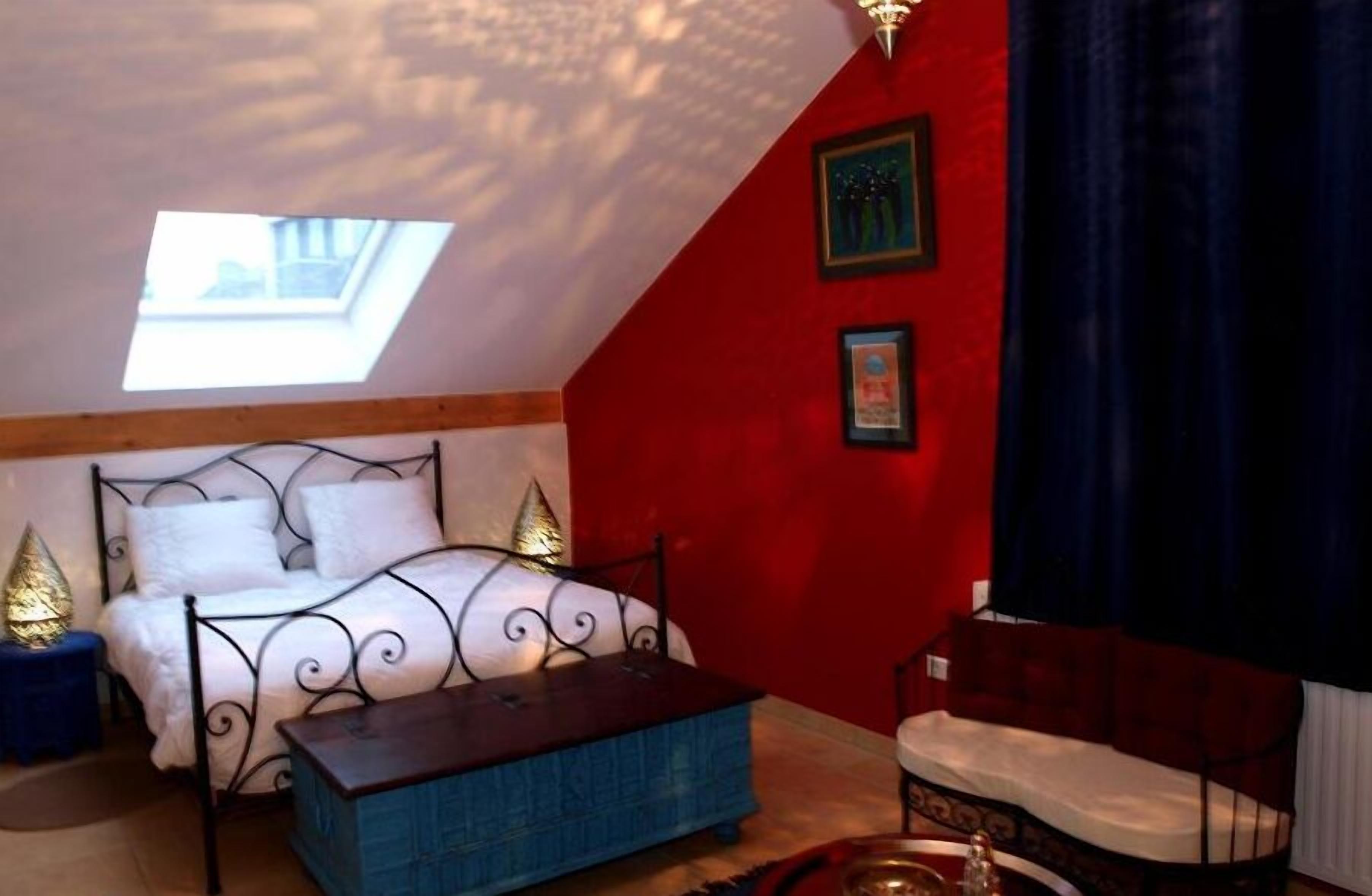 Ein Schlafzimmer mit schrägen Wänden, einem Oberlicht, einem Bett mit Metallgestell, einem blauen Pouf und einer roten Akzentwand.