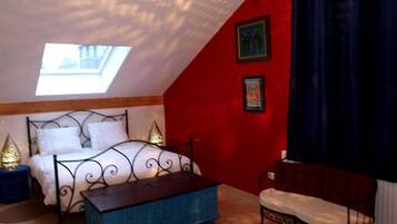 Ein Schlafzimmer mit schrägen Wänden, einem Oberlicht, einem Bett mit Metallgestell, einem blauen Pouf und einer roten Akzentwand.